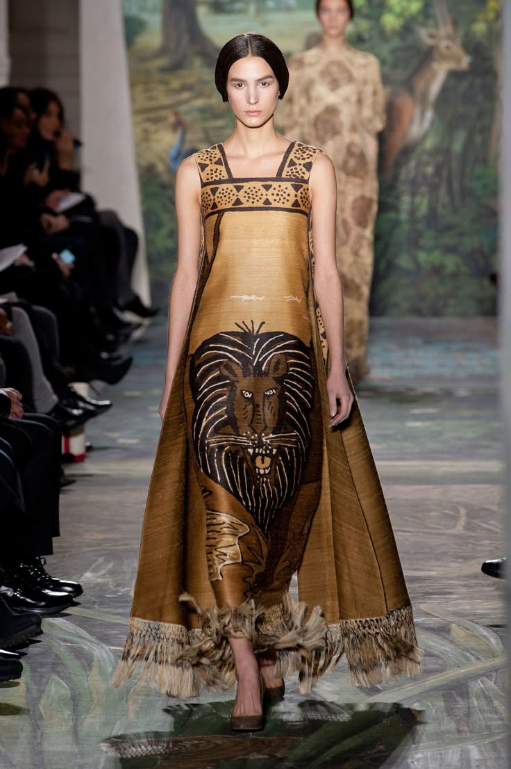 a model walks down a runway wearing a brown sleeveless dress with a lion print on the front. the dress has fringe at the bottom. an audience is seated on the left side of the image, and a jungle scene is in the background. another model is standing behind the main model, wearing a patterned dress.