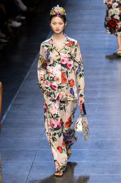 a model walks down a runway wearing a floral printed pajamastyle set. the outfit consists of a longsleeved shirt and wideleg pants in a delicate beige base with a vibrant floral pattern featuring roses and birds. the model also wears a jeweled crown and carries a small embellished handbag. the overall style is luxurious and feminine, evoking a sense of spring or summer fashion. the background suggests a professional fashion show setting.