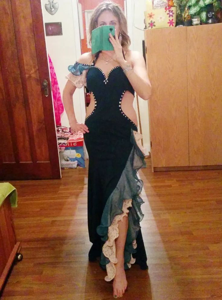 a woman stands barefoot in front of a mirror, taking a selfie with a green tablet that obscures her face. she's wearing a long, black evening gown with cutouts along the waist and studded detailing. the dress features teal and white ruffles cascading down one side. the background shows a wooden door, a cabinet, and items stacked behind her. the floor is hardwood.