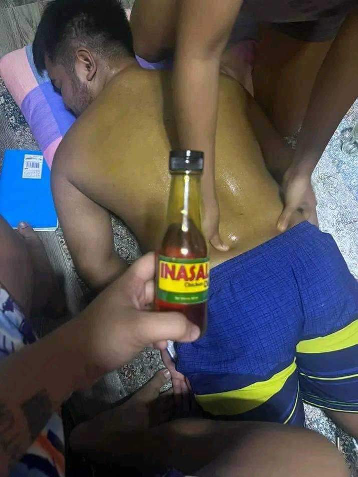 a man lies face down on the floor while two other people massage his back. he is shirtless and wearing blue and yellow shorts.  one of the people holds a bottle of inasal, a filipino condiment typically made with chicken fat and annatto, implying it's being used for the massage.  the scene takes place indoors on a floor with a blue pillow and notebook visible. the lighting is artificial and the perspective is from above.