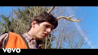 a man with brown hair and a beard is wearing a plaid shirt, an orange vest, and deer antlers on his head. he is smiling and looking at the camera. the background is a forest with trees and branches. the vevo logo is visible in the bottom left corner.