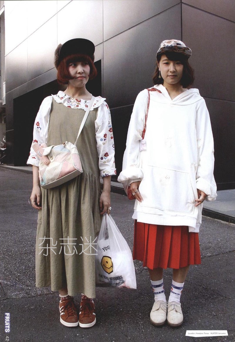 the image features two young women showcasing distinct harajuku street styles.  the woman on the left wears a long khaki dress over a floral blouse, paired with a brown bucket hat and sneakers. she carries a colorful patchwork bag.  the woman on the right sports an oversized white hoodie layered over a red pleated skirt, accessorized with a patterned cap, white socks, and brown shoes. she holds a plastic shopping bag.  both women pose against a backdrop of a modern building facade with a muted gray and silver color scheme. the overall atmosphere is casual and stylish, capturing the essence of japanese street fashion.