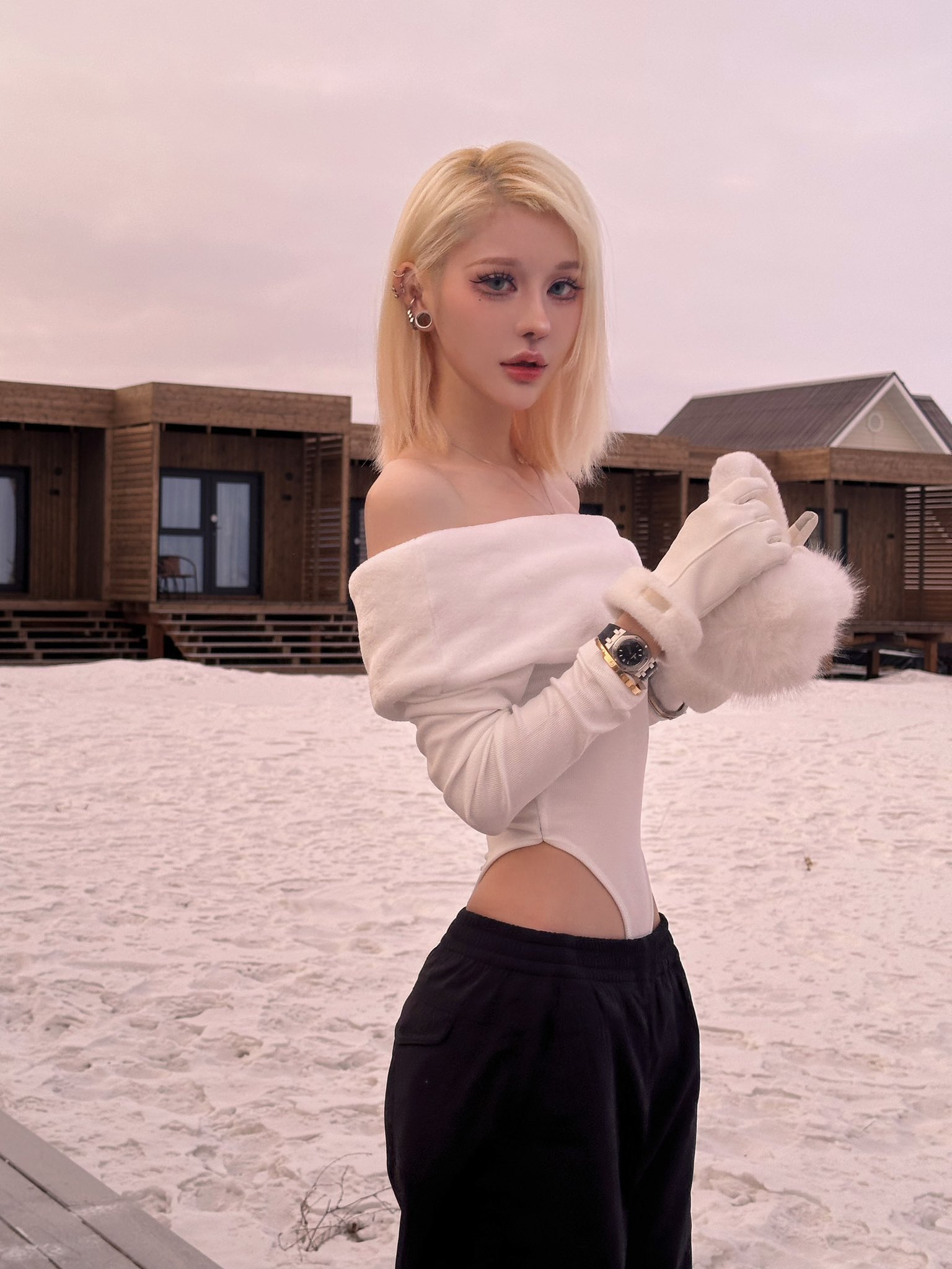 a young blonde woman in a stylish white offshoulder bodysuit and black pants poses outdoors in a snowy setting. she is wearing fur gloves and a watch.  the background features modern wooden buildings. the overall mood is elegant and cold.