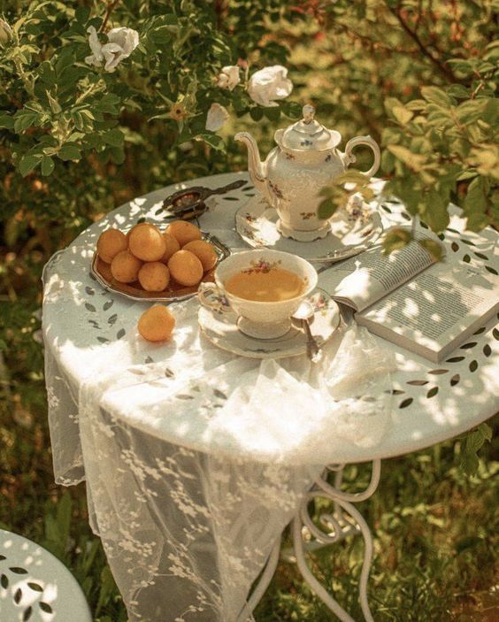 the image shows a white table with a lace tablecloth, set outdoors in a garden.  sunlight filters through the leaves of the surrounding trees and shines on the table.  a vintage teapot and teacup are arranged on the table, next to a plate of apricots.  a book lies open on the table beside the teacup.