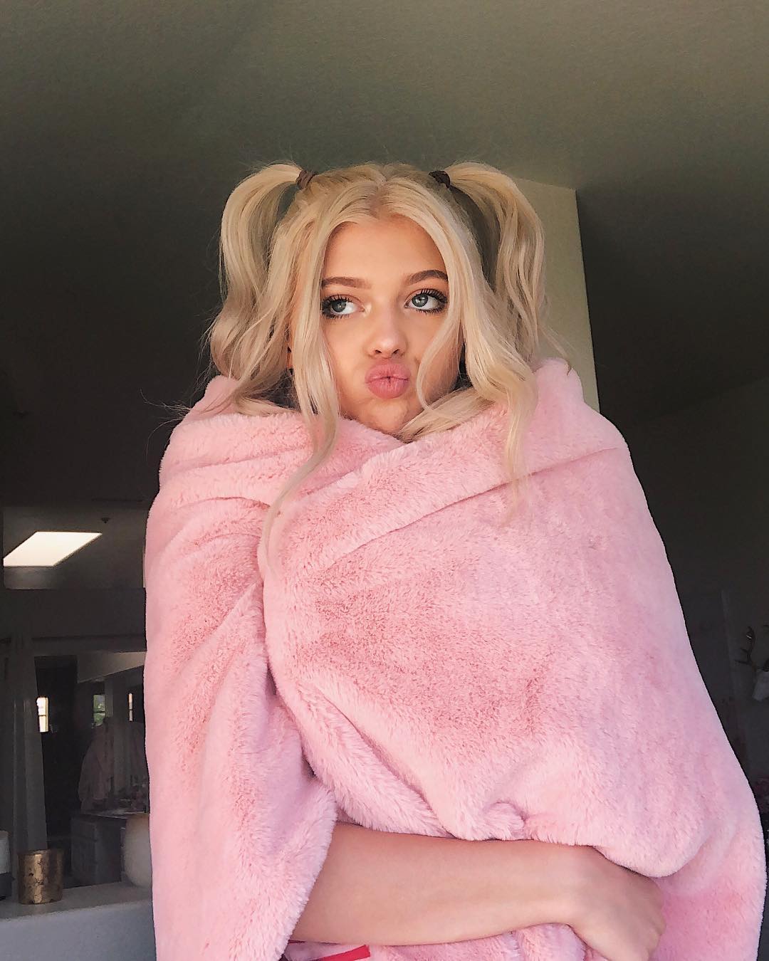 a young woman with blonde hair styled in pigtails, blue eyes, and puckered lips is wrapped in a pink, fluffy blanket. she is indoors and appears to be posing for the photo. the overall impression is youthful, cute, and cozy.