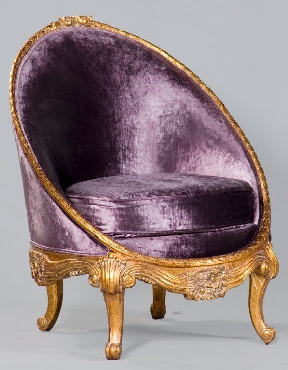 a luxurious chair with a rounded back upholstered in purple velvet. the chair has ornate gold trim around the edges and along the base. it has four legs with curved feet. a square purple cushion sits on the seat. the background is a neutral gray.