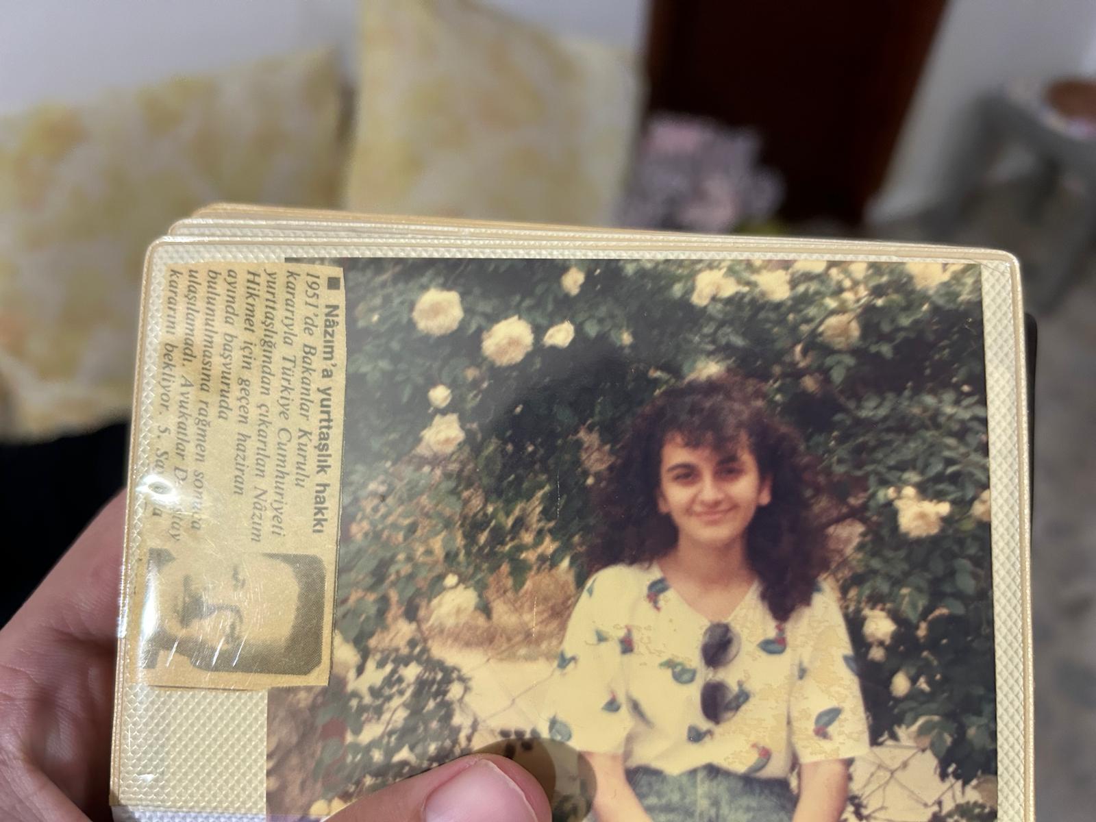 a closeup shot of a vintage photograph shows a young woman with curly brown hair smiling gently. she's wearing a lightcolored, shortsleeved shirt with a floral pattern and is standing in front of a lush rose bush filled with white and light pink roses. the photo is slightly faded and has a vintage feel. the photo is part of an album and there's a piece of paper visible in the corner with text in turkish, possibly relating to  nazım hikmet.  a hand is visible holding the photo album in the lower left corner.
