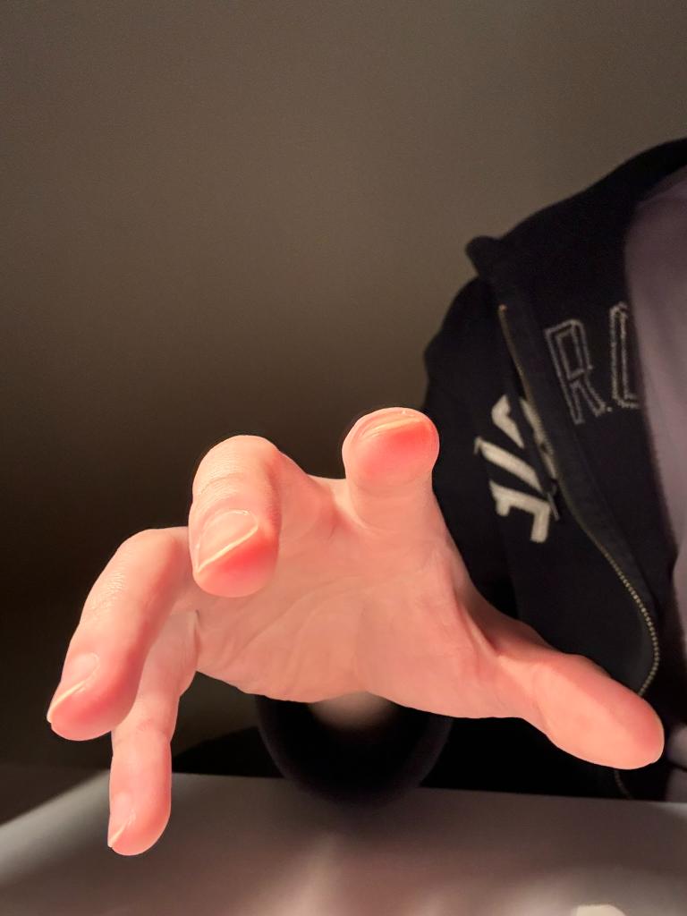 the image shows a closeup of a hand reaching forward, with the fingers spread out. the hand is lit from above, creating highlights on the knuckles and fingertips. the hand appears to be reaching towards something out of frame.