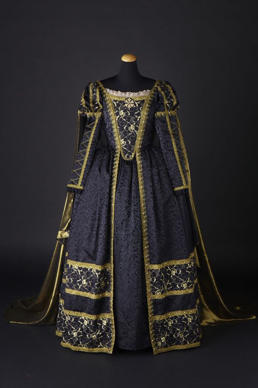 a fulllength photograph shows a dark blue and gold gown displayed on a mannequin, likely in a medieval or renaissance style. the gown is predominantly dark blue with intricate gold trim detailing, including decorative panels on the bodice, sleeves, and the lower portion of the dress. gold fabric drapes from the sleeves, adding a sense of grandeur and movement to the design. the mannequin is placed against a dark background, allowing the gown to stand out as the focal point of the image.