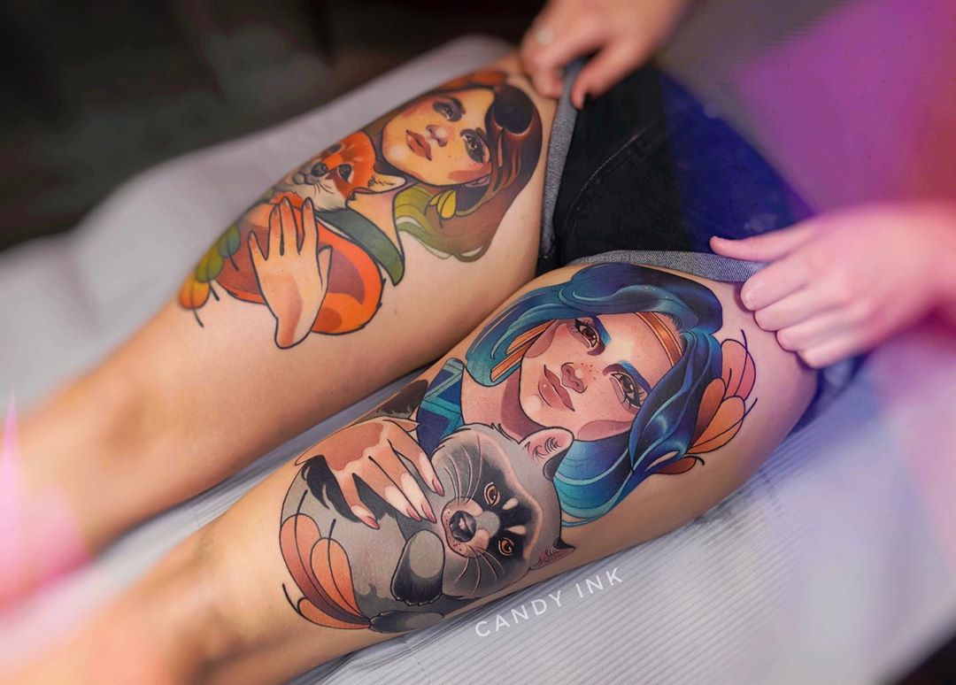 the image shows a pair of women's legs with two large tattoos on the thighs. the tattoo on the left thigh depicts a woman with long brown hair holding a red fox. the tattoo on the right thigh depicts a woman with blue hair holding a raccoon. the tattoos are in a colorful, illustrative style. the background is a white sheet.