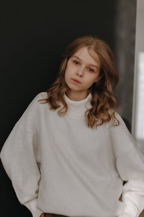 a portrait of a young woman with blond, wavy hair wearing a white, longsleeved turtleneck sweater against a dark background. she has a casual and slightly pensive expression.