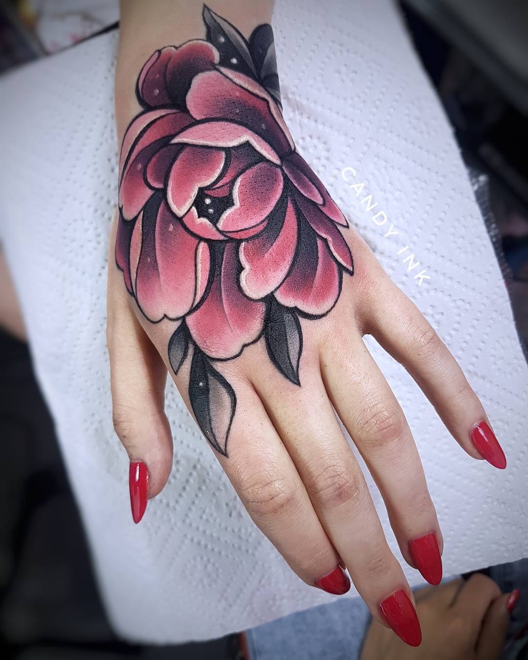 a woman's hand with a fresh neotraditional style tattoo of a pink peony flower. the tattoo is on the top of the hand and covers most of the surface area. the tattoo has been applied with black ink and a variety of pink and red inks. the woman has long red nails. the background is white.