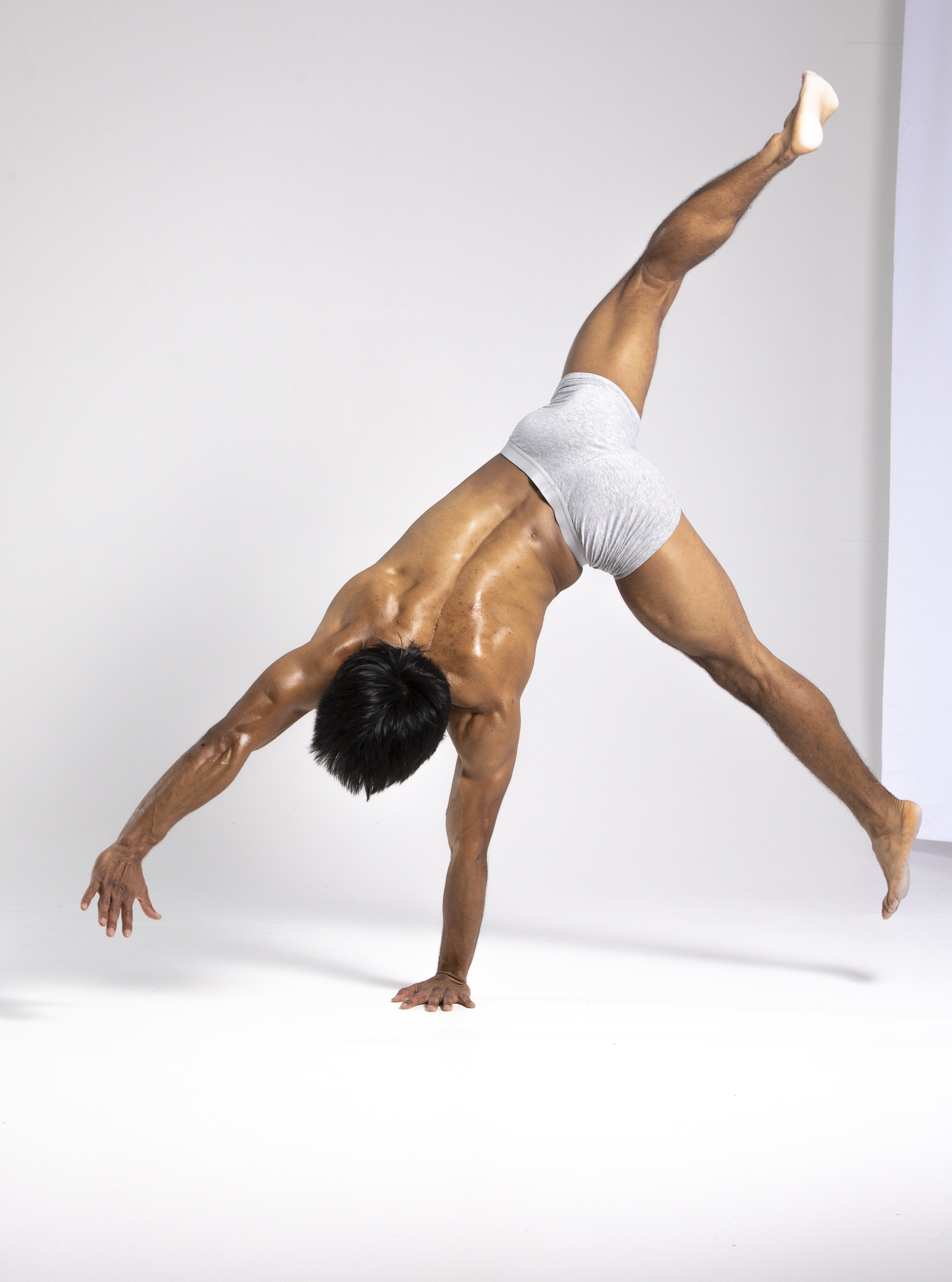 a muscular, barechested man in gray underwear is performing a handstand in a studio. his body is oiled or sweaty, highlighting his physique. he's balanced on one hand while the other arm is extended. one leg is raised straight up, and the other is extended to the side, showcasing his flexibility and strength. the background is a simple white backdrop, putting the focus on the man's athletic performance.