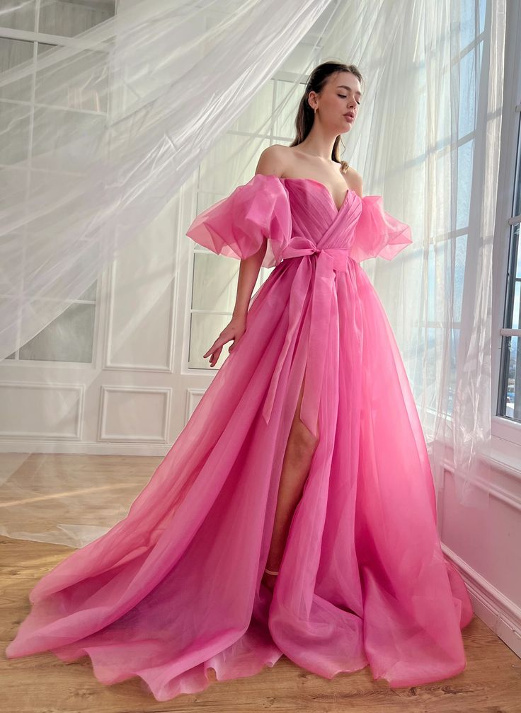 a beautiful woman is posing in a pink dress. she has on a long, flowy dress with a sweetheart neckline and offtheshoulder straps. she has long dark hair and is wearing minimal makeup.