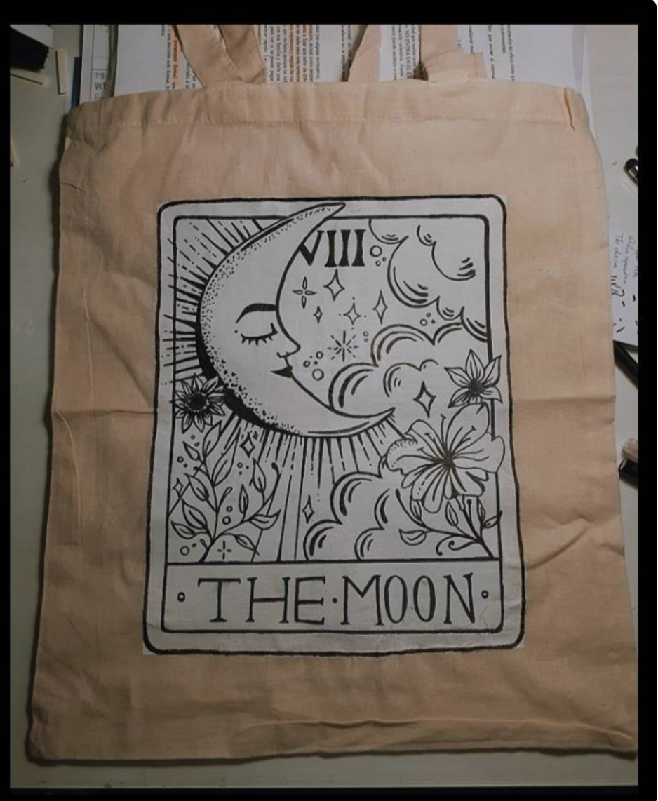 a plain tote bag lays flat on a surface. the tote bag is a light tan color and features a blackandwhite drawing of "the moon" tarot card.  the drawing features a crescent moon with a face, flowers, and stars, with the words "the moon" at the bottom.