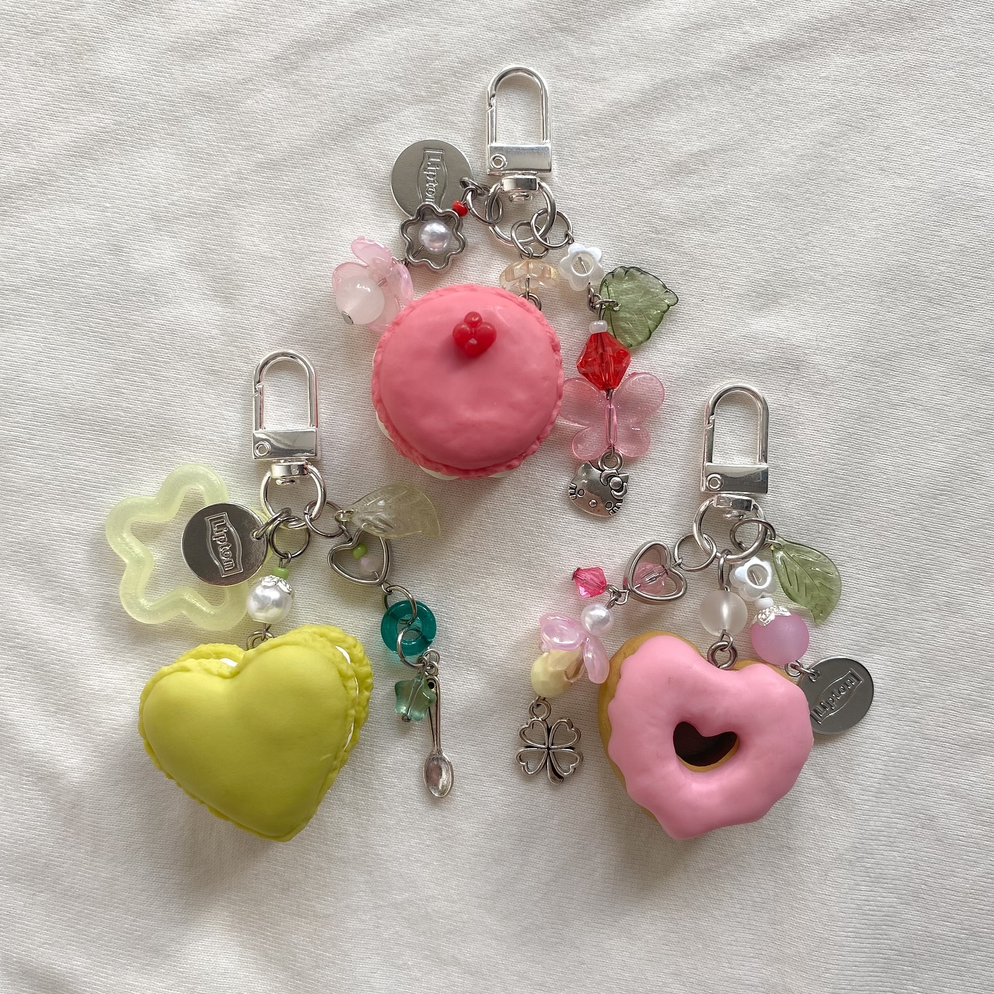 three decorative keychains are arranged on a textured white surface. each keychain features a different dessertthemed pendant attached to a silver clasp with a variety of beaded charms and trinkets. the keychains are arranged in a row, showcasing the different pastelcolored desserts and the diverse collection of charms and trinkets attached to them.