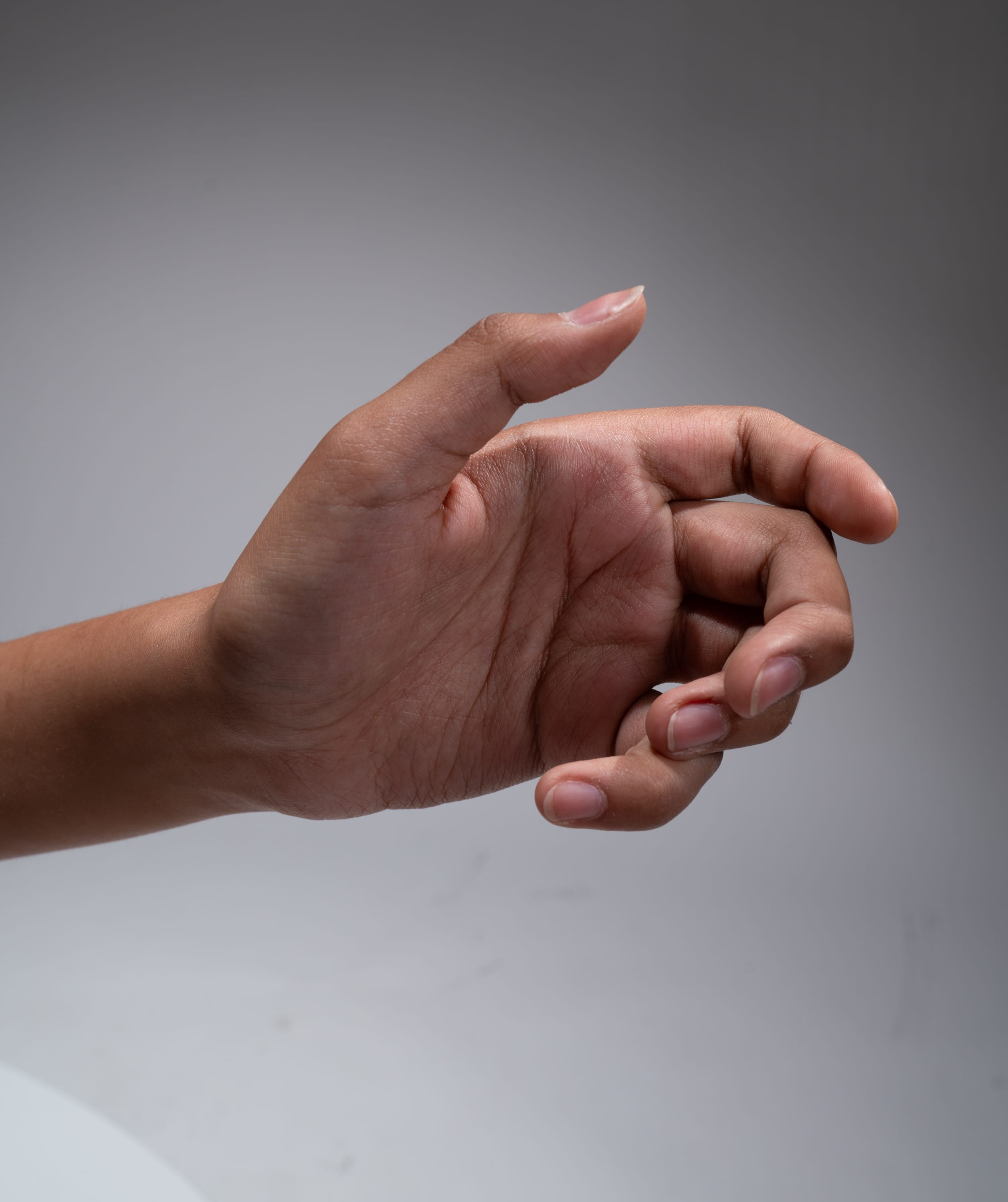 a hand is extended with the palm up, fingers slightly curled, against a gray background. the hand belongs to a person with darker skin, and the image is lit by studio lighting, offering a clear side view of the hand.