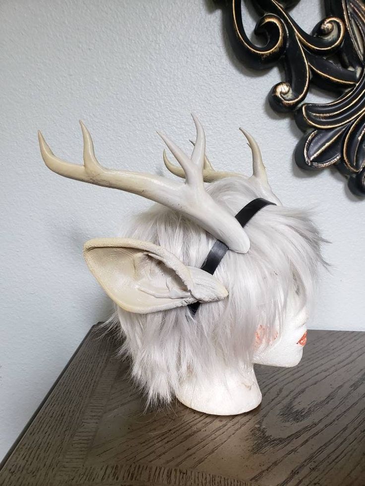 a white deer antler headband sits on a mannequin head with a white wig. the antlers are made of plastic or resin. the headband is black with a white antler on each side. the mannequin head is turned to the side, showing the antlers. the mannequin is sitting on a wood table. the photo is taken in a room with a white wall and a black ornate mirror in the background.