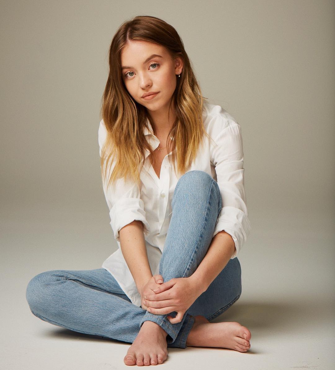 a young woman with long blonde hair sits barefoot on a light beige background. she is wearing light blue jeans and a white buttondown shirt. her expression is relaxed and she looks directly at the camera.  the image is a professional portrait, likely for a modeling portfolio or similar purpose.