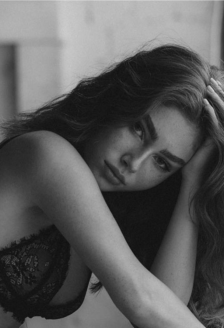 the image is a black and white, closeup portrait of a young woman. she has long, wavy brunette hair and noticeable freckles. she is wearing a black lace bra and her expression is pensive and somewhat melancholic. one arm is resting on her head, and the other arm is draped across her body. the lighting is soft and diffused, creating a moody and intimate atmosphere. the overall feel is artistic and sensual.