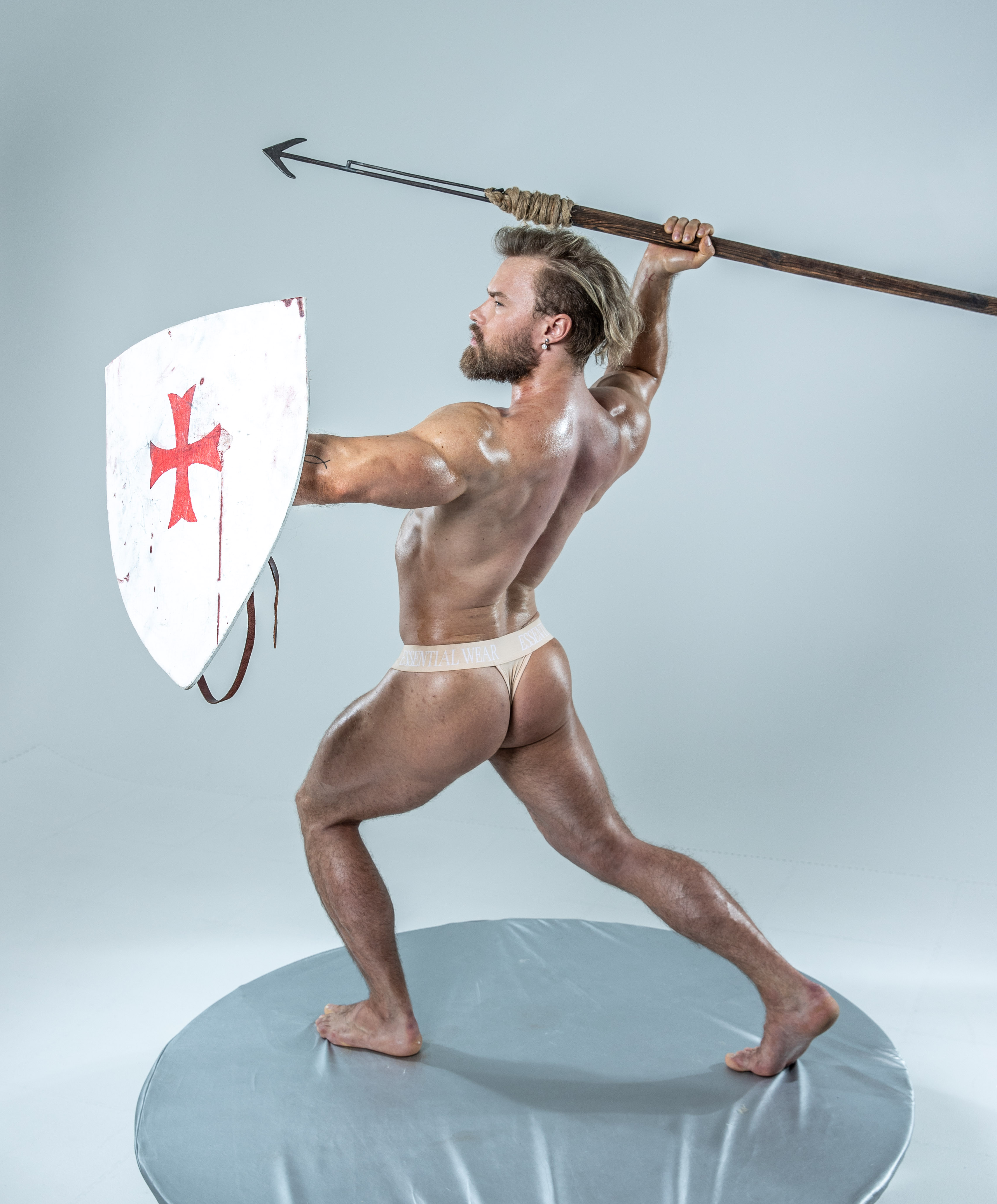 a muscular man, wearing only a loincloth, poses dynamically against a light grey background. he holds a wooden spear aloft in his right hand and a large, round shield adorned with a red cross in his left hand. the image evokes classical imagery of warriors and gladiators. his physique is welldefined, highlighting his strength and athleticism. the studio setting and bright lighting emphasize the subject and his powerful stance.