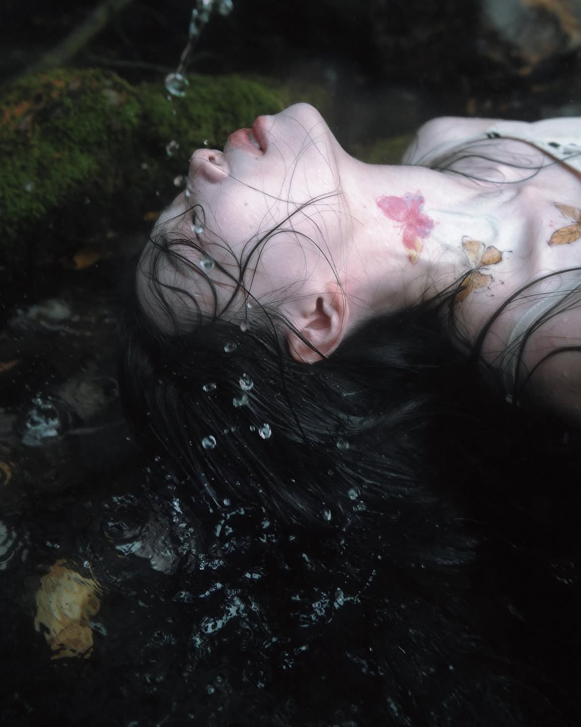 a fairskinned woman with dark, wet hair lies in what appears to be a shallow pool of water. water droplets are visible on her face and hair. her head is tilted back, and a mosscovered object is visible to the left. the woman has butterfly tattoos on her neck and collarbone. the overall image has a surreal, ethereal, and somewhat melancholic feel.