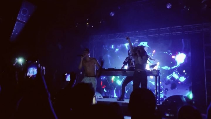 a photo of a performer singing with a microphone, shirtless, on a stage with two other people, one standing at a dj turntable and the other raising her hand in the air. the audience is facing the stage, illuminated by various lights and the glow of cell phones.