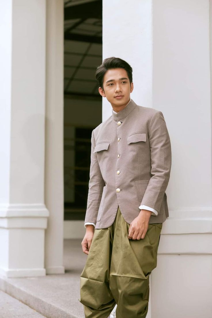 a young man in a traditional thai outfit stands in front of a white wall. he is wearing a brown jacket and green pants and is looking off to the side. he is smiling slightly. the background is blurred.