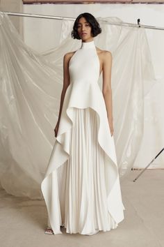 a woman in a stylish white dress, standing in a studio, against a white background. she is posing elegantly, with her hair pulled back. the dress is a highneck style, with a flowing skirt and an intricate design that enhances the overall elegance of the outfit.