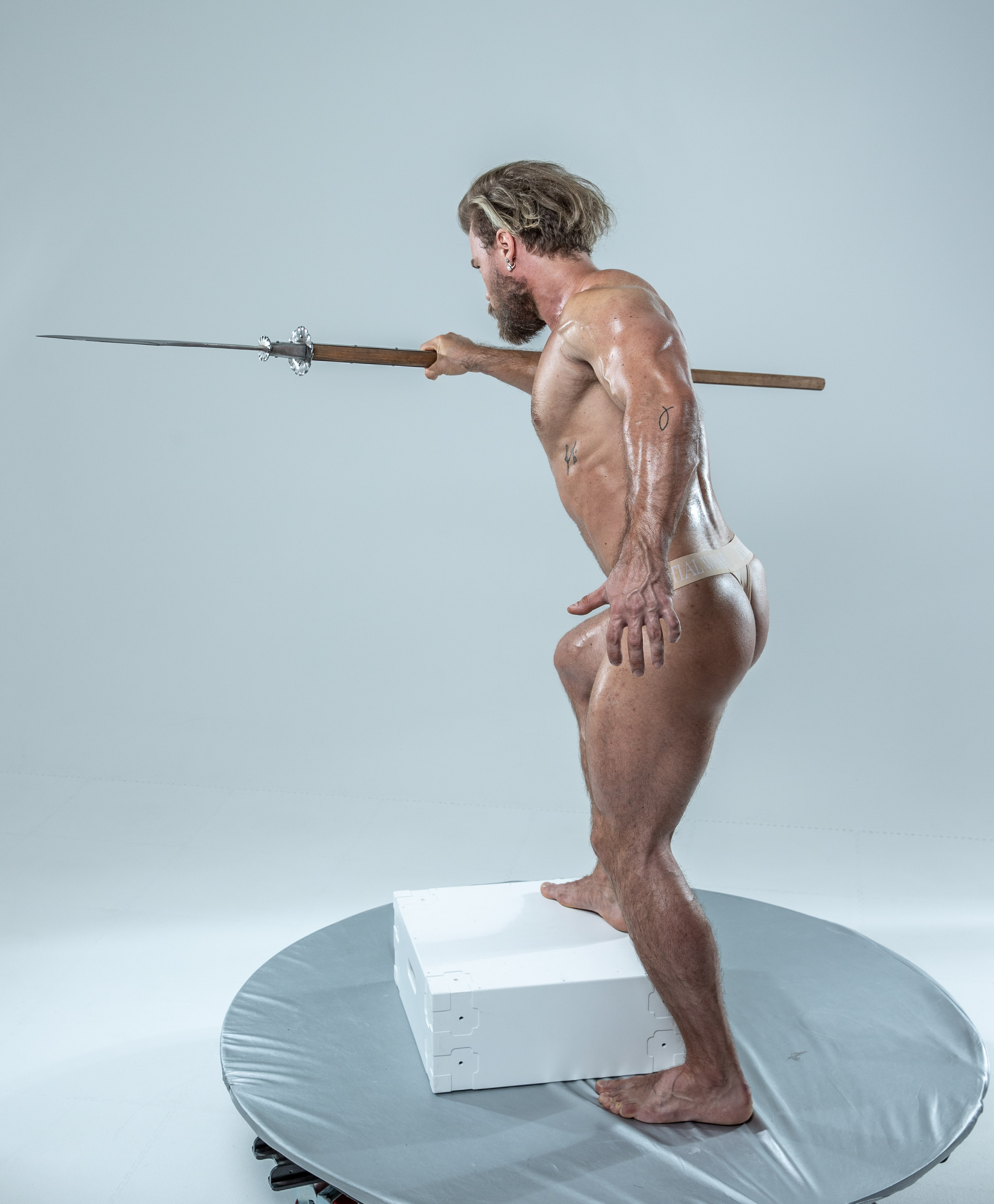 a muscular, adult man with long blond hair pulled back stands balanced on a white box. he is holding a spear in both hands, pointing it to the left.  the spear is level with his shoulders. he has a thin white band around his upper thighs. the background is a light gray.  he is positioned on a round, gray platform.