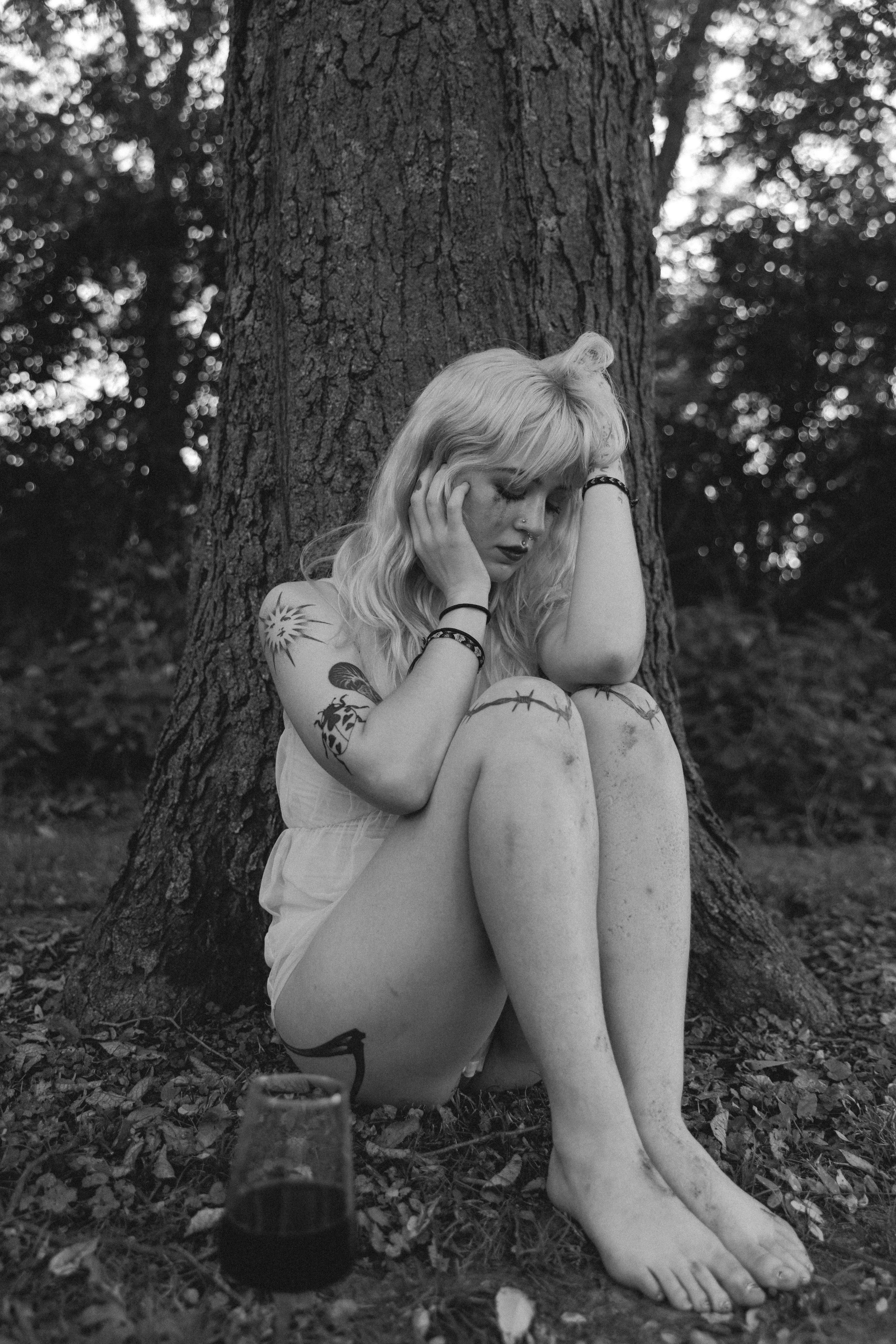 a black and white photograph of a young woman sitting against a large tree in a forest. she is wearing a short white dress and has tattoos on her arms and legs. she is hugging her knees and has her head bowed down. she appears sad or contemplative. the photo has a dark, moody atmosphere.