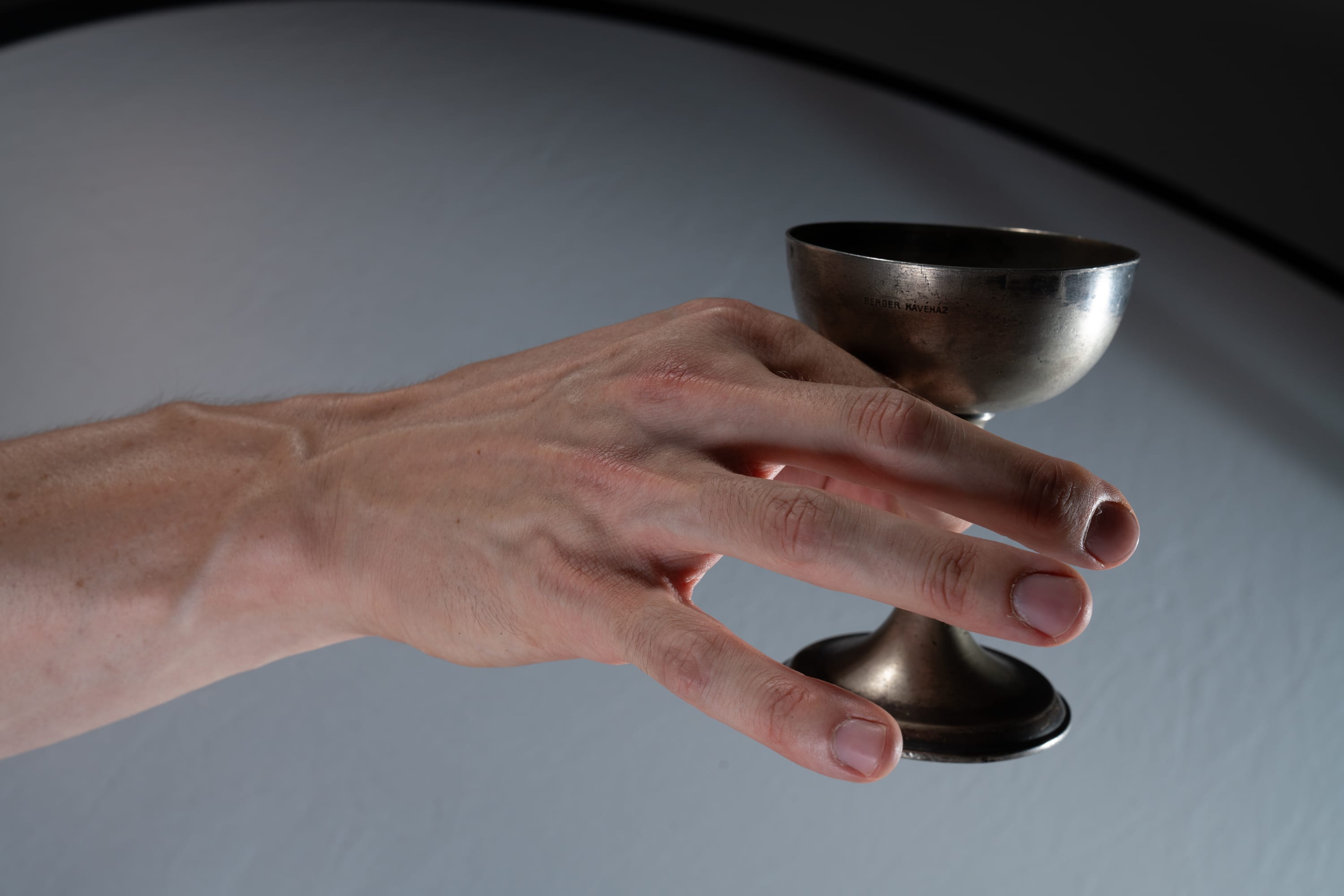 a hand holding a silver cup against a white and black background. the hand is extended with the cup delicately balanced on the fingers.  the lighting emphasizes the details of the hand and the reflective surface of the cup.