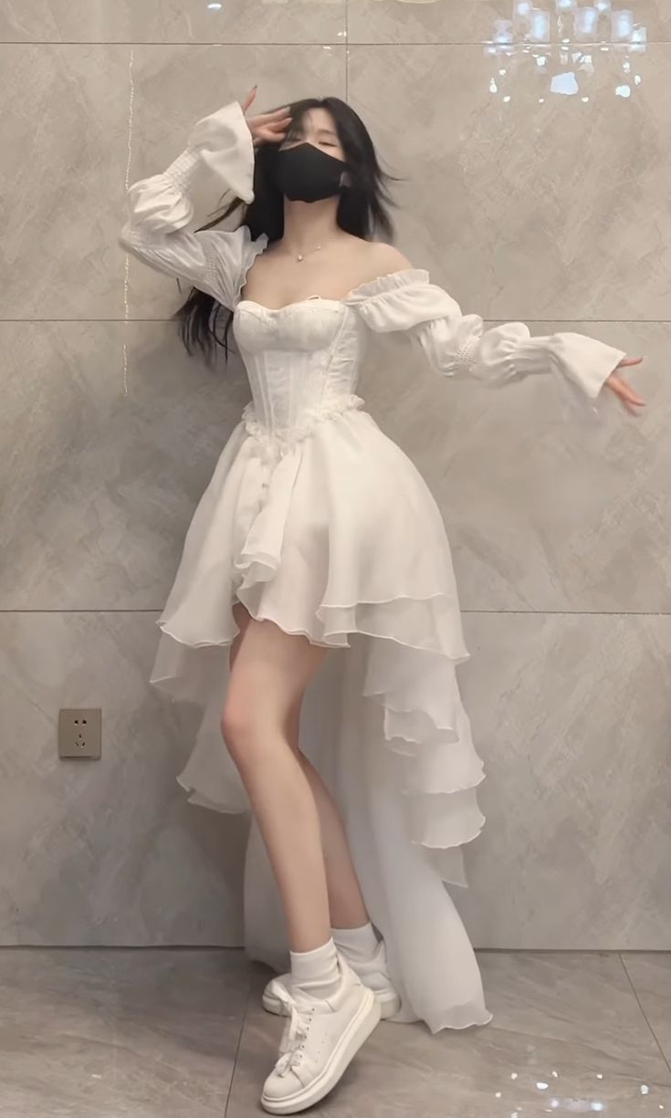 a young woman with long dark hair is posing in a white highlow dress. the dress has puff sleeves and a corset bodice. she is wearing white sneakers and socks. she is also wearing a black face mask. the background is a light beige wall with a subtle pattern. the overall style is elegant, minimalist, and chic.