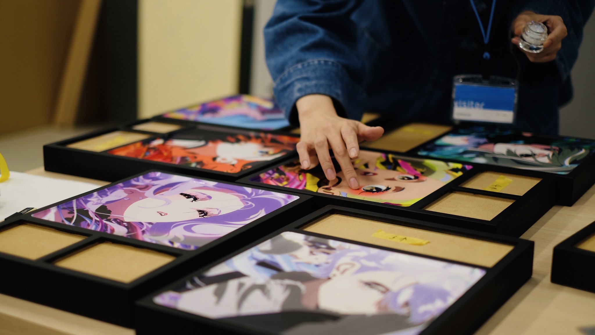 the image showcases a table filled with framed animestyle artwork. an artist's hand is seen arranging one of the prints. the prints feature vibrant colors and characters with large eyes, a hallmark of the anime and manga style. the setting suggests an art fair or convention, likely an artist alley where independent artists sell their work.