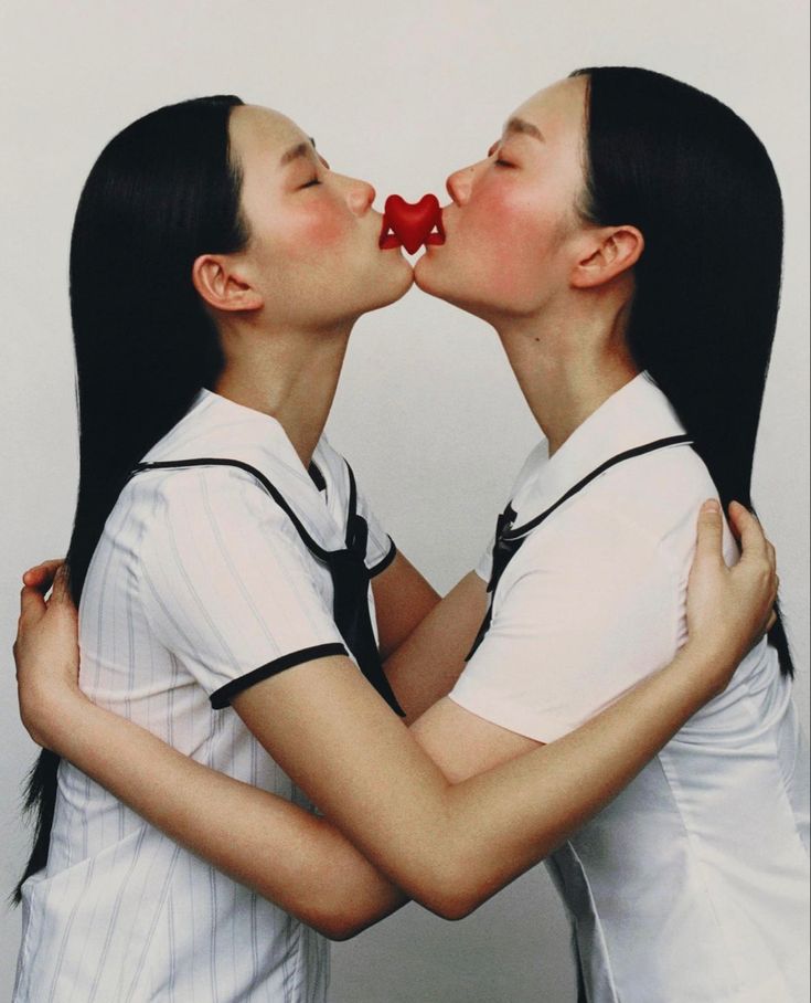 the image shows two young asian women, possibly twins, who are embracing and appearing to kiss. a small, red heartshaped object is positioned between their lips. they are wearing matching white school uniforms with black sailor collars. their arms are intertwined, and the overall mood is one of affection and tenderness. the background is simple and uncluttered, focusing attention on the girls. the photograph is brightly lit, highlighting their youthful and soft features.