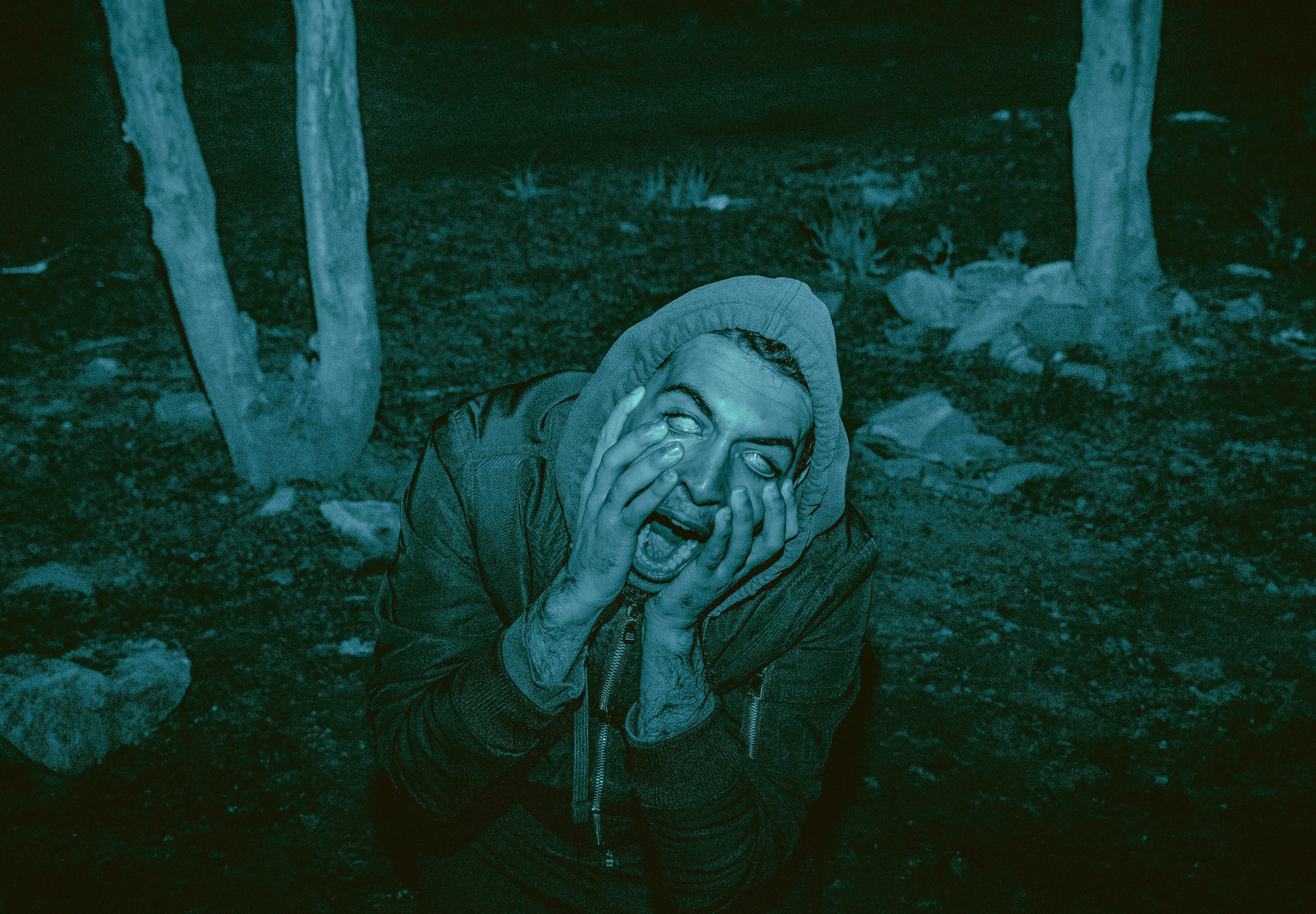 a black and white photo of a man dressed as a zombie. his face is painted white, and he has black around his eyes. he is wearing a hooded jacket and is making a scared face. the photo is tinted bluegreen.