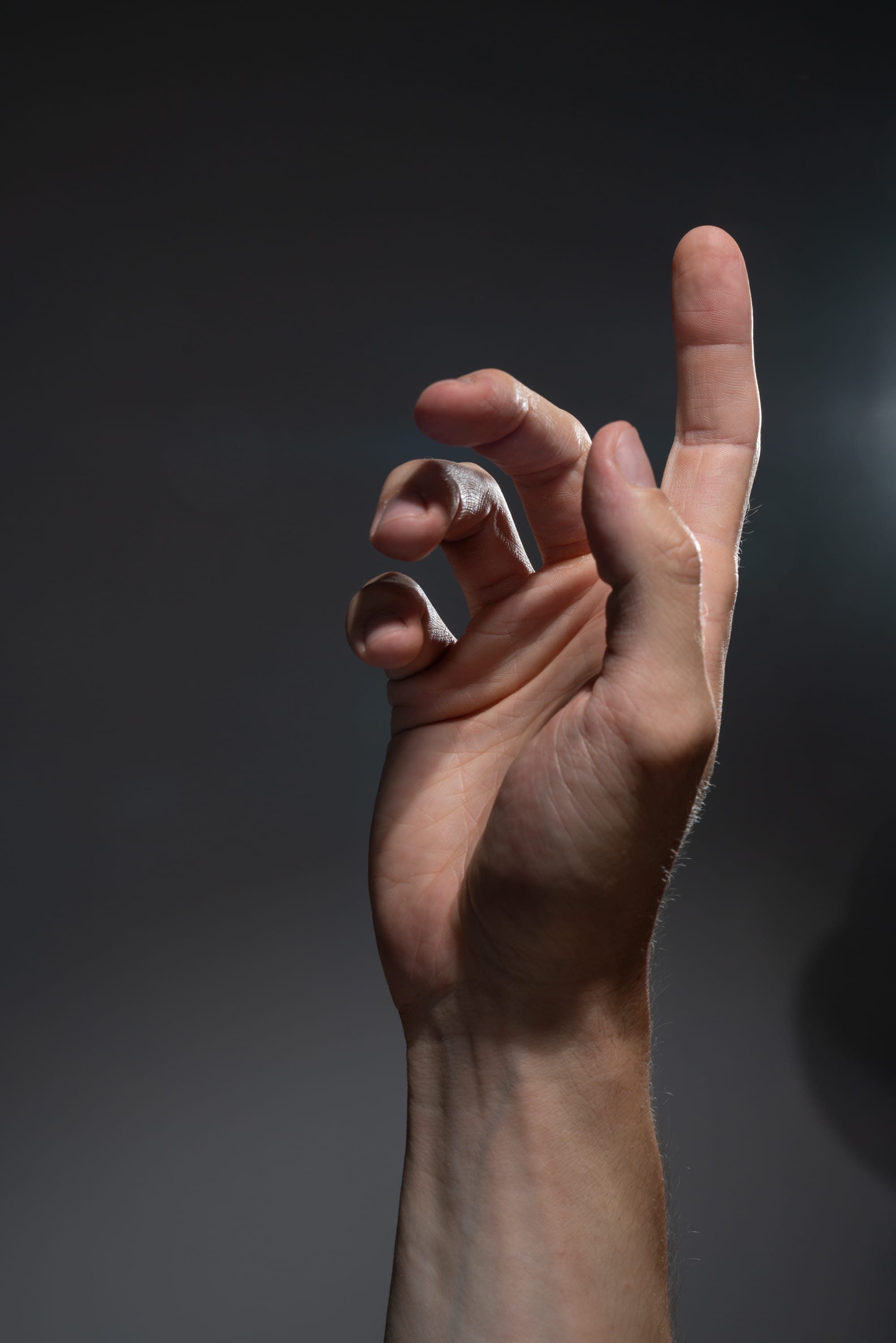 a closeup of a man's right hand extended against a gray background. the hand is illuminated by a spotlight, highlighting the details of the skin and fingers. the fingers are slightly curled inward, creating a sense of tension and anticipation.