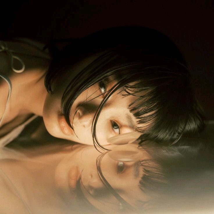 a woman with short black hair lies on her side with her face reflected in a mirror. the lighting is soft and warm, creating a dreamy atmosphere.