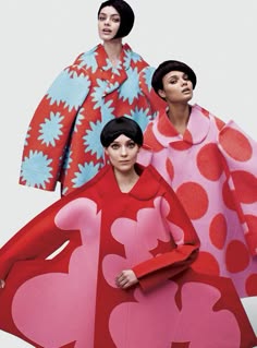 three women are dressed in colorful coats posing for a photoshoot. the woman in the front is wearing a red and pink coat with pink polka dots on the skirt. the woman in the middle is wearing a pink coat with red polka dots and the woman on the right is wearing a light blue and red coat. the woman in the front has her hands on her hips, the woman in the middle has her hands on her waist, and the woman on the right has her hands on her chest. the women are all looking at the camera.