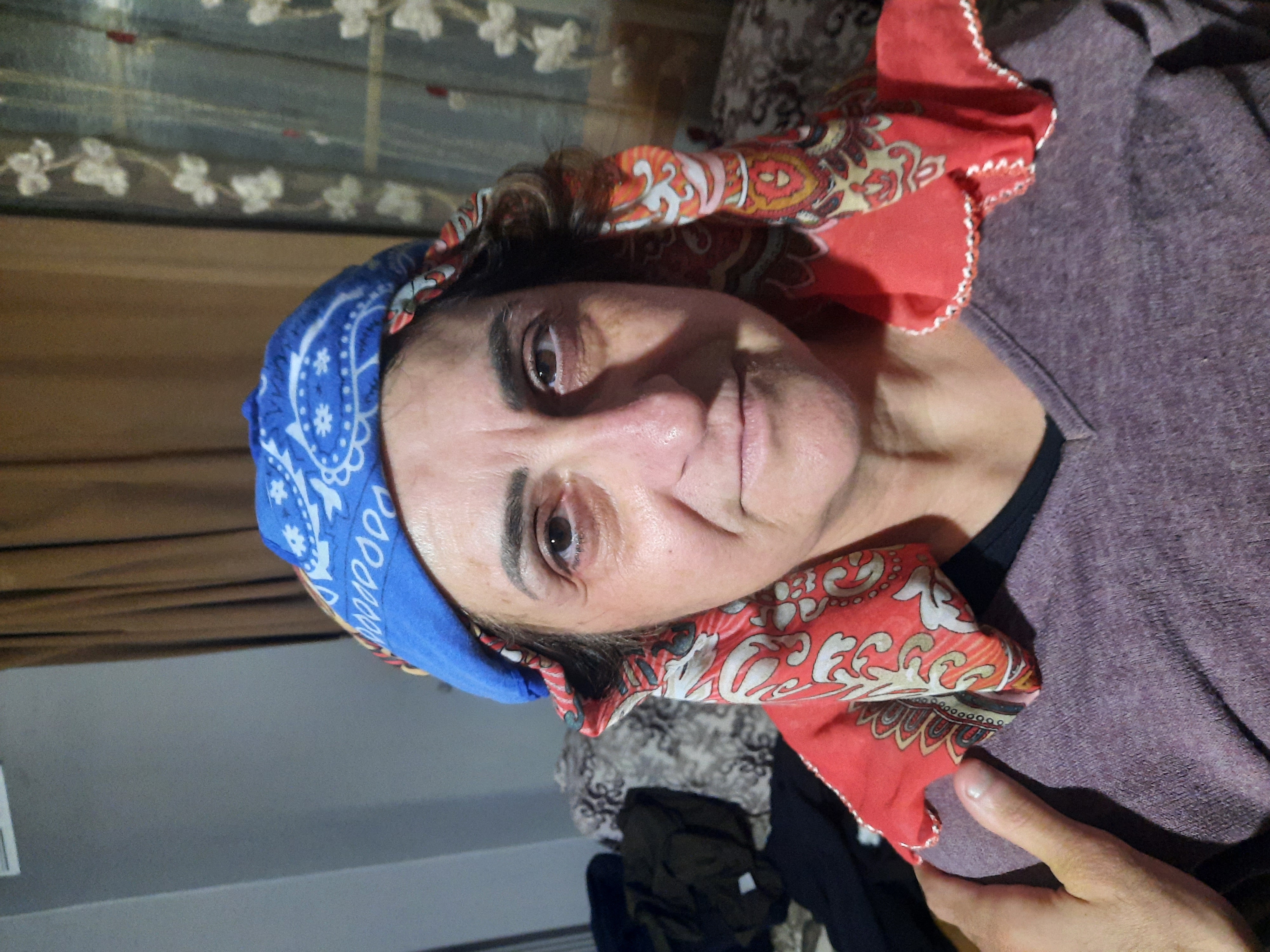 a closeup portrait of a woman wearing a blue bandana and a red patterned headscarf. a hand rests on her shoulder. the background includes a curtain and a window.