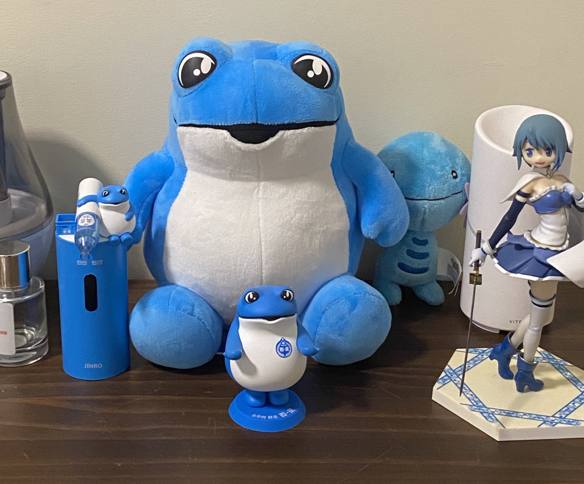 a blue frog plush toy is sitting on a wooden table. there is a smaller blue frog figurine sitting in front of it. a blue anime girl doll with white and blue clothing is standing on the table to the right of the plush toys. there is a tall blue and white container behind the girl doll and a smaller blue plush frog toy behind the large plush frog. the background is a light tan wall.