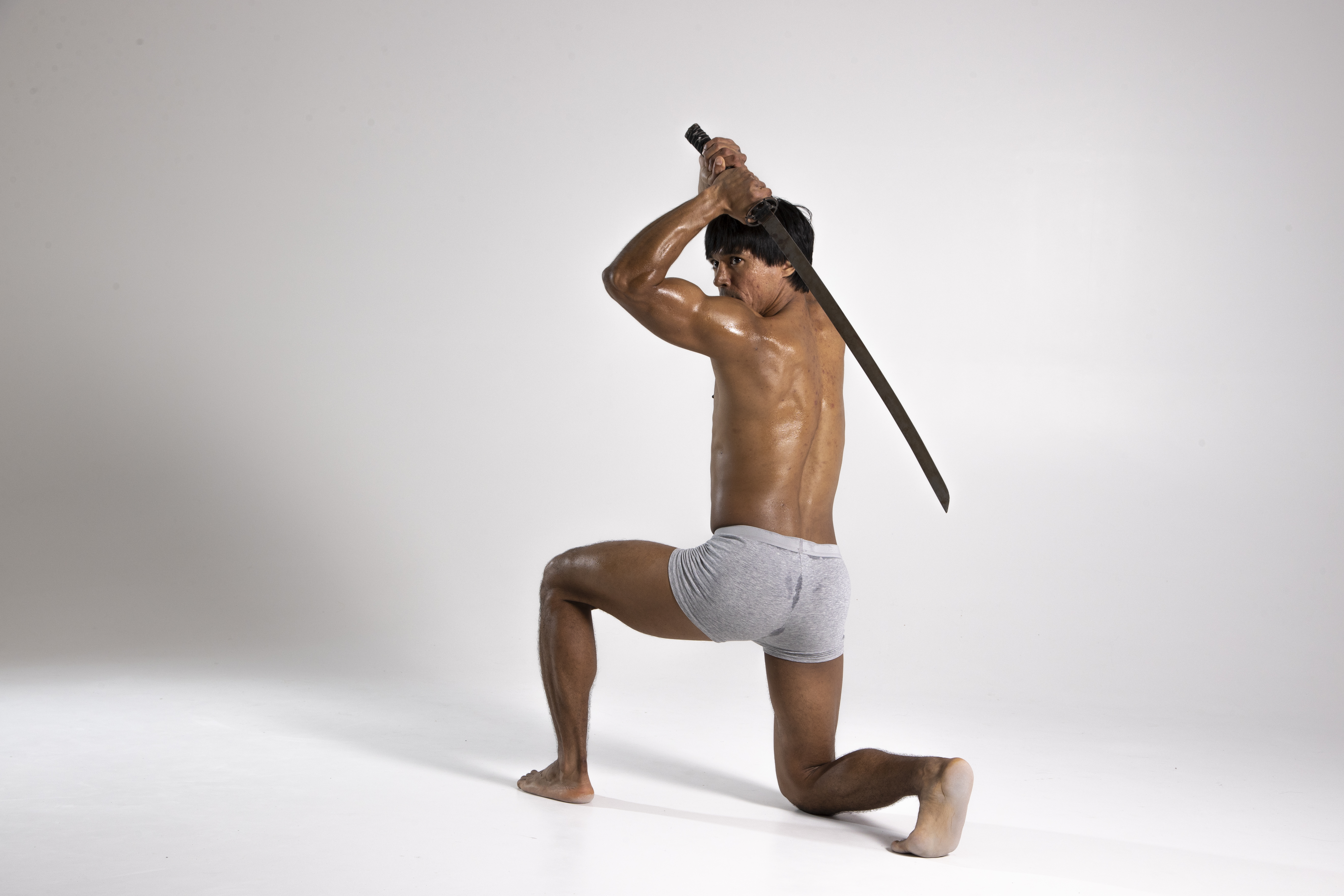 a muscular, shirtless asian man is kneeling in a studio with a gray background, holding a sword above his head. he is wearing gray underwear and is sweating, suggesting a workout. he is looking back over his shoulder with a determined expression. the sword is large and appears to be made of dark metal. his toned body and the dynamic pose convey strength and athleticism.