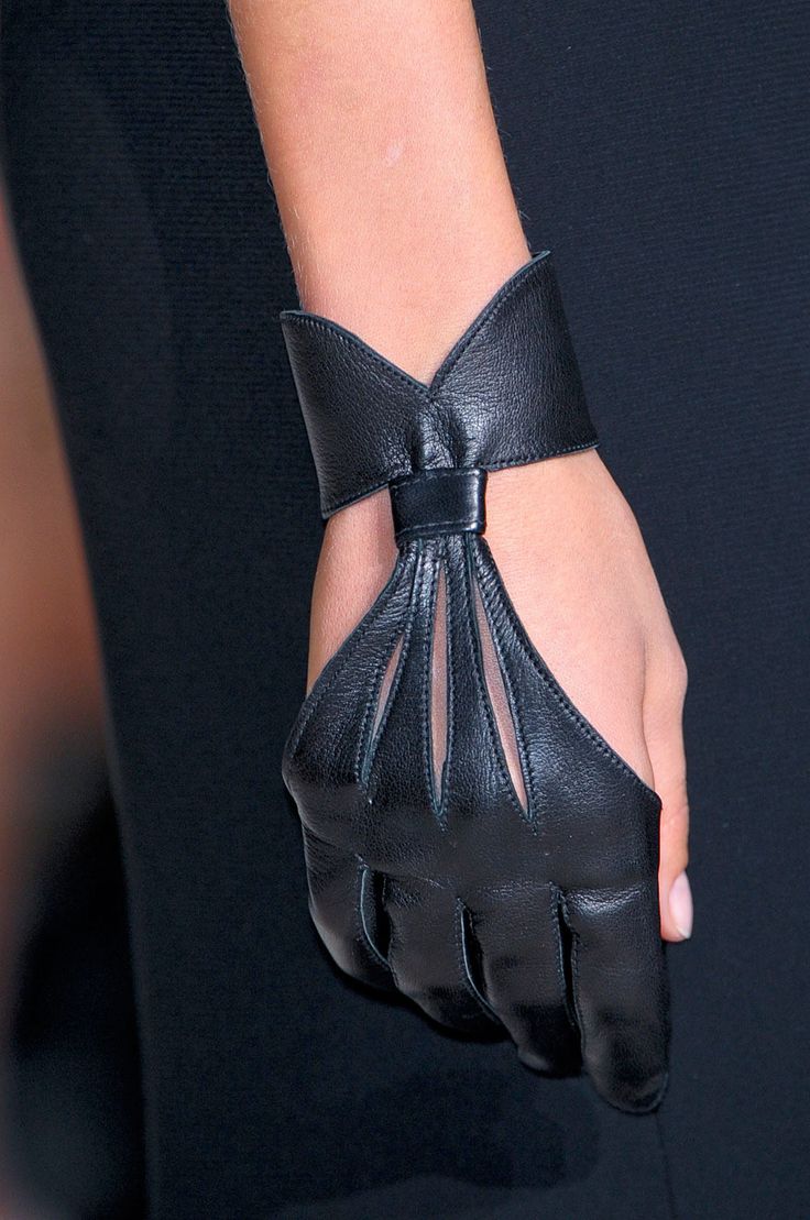 a closeup of a woman's hand wearing black leather gloves. the gloves are cut out with a sheer material, revealing the woman's skin. the gloves are styled with a cuff at the wrist, which is also made of leather. the woman is wearing a black dress and the gloves are a stylish and elegant accessory.
