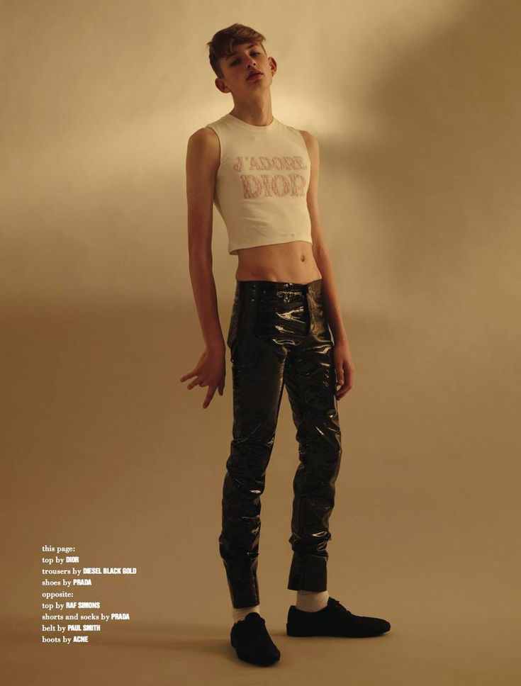 a young man poses for a fashion shoot in a studio setting. he is wearing a white crop top with the words "j'adore dior" on it, black pants, and black shoes. he has short brown hair and is looking at the camera. the background is a solid beige color. the image is welllit and the focus is on the model.