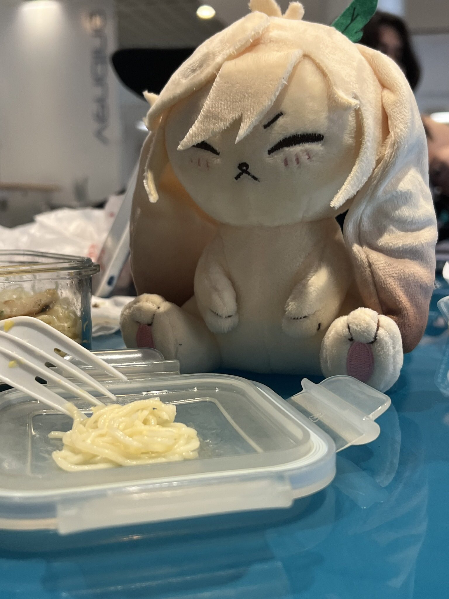 a white bunny plush toy sits on a blue table next to a white plastic container with noodles inside. a fork is sticking out of the container.