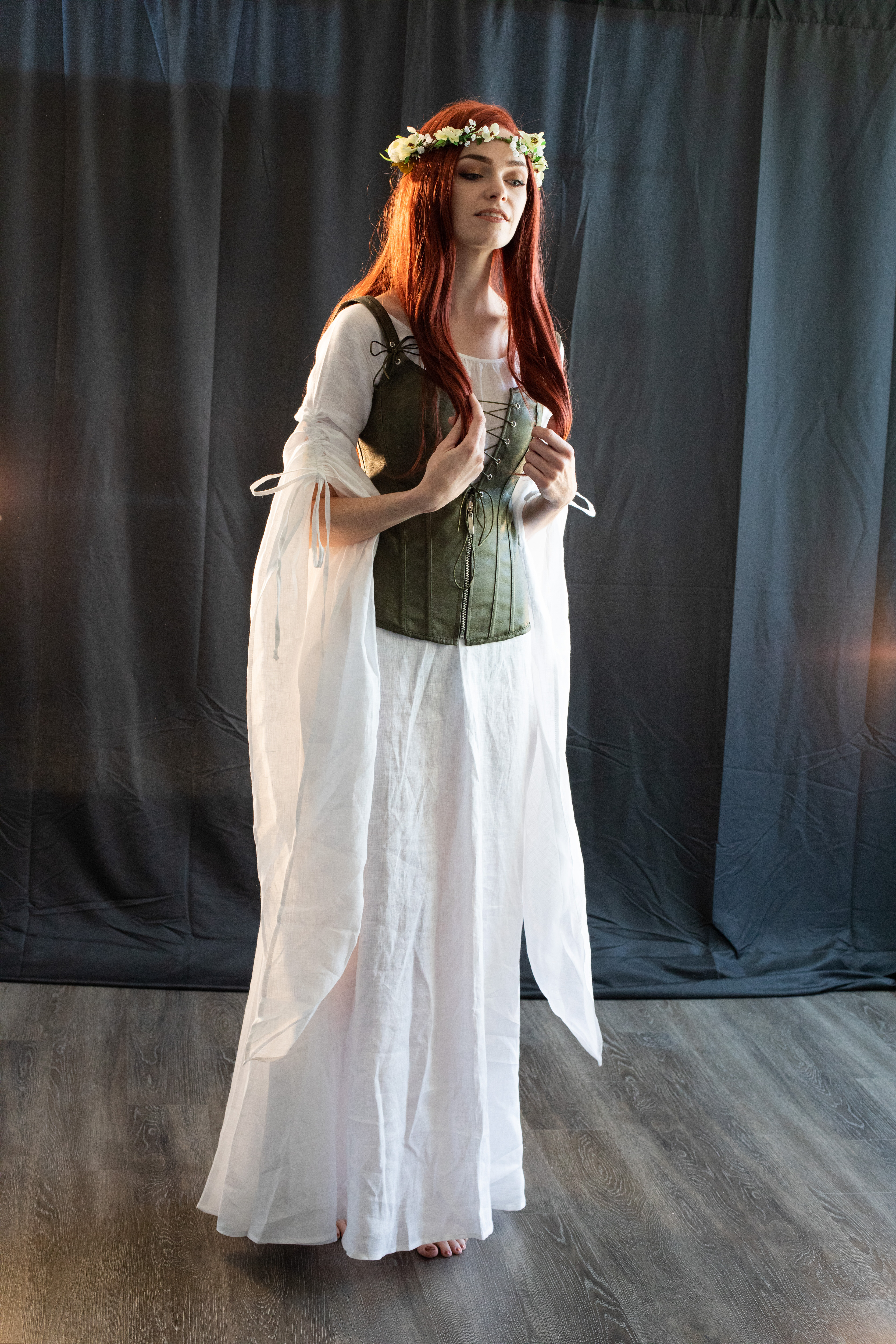 a young woman with long, red hair stands in a studio setting. she wears a white medievalstyle dress with flowing sleeves and a green corset. a delicate flower crown adorns her head. the background is a dark, textured curtain, and the lighting is soft, creating a dreamy atmosphere. her pose suggests she might be adjusting her corset.