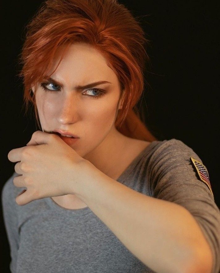 a closeup portrait of a woman with red hair looking away from the camera, wearing a grey shirt with an american flag patch. her left hand is covering her mouth, and she appears serious and focused. the background is blurred and dark.