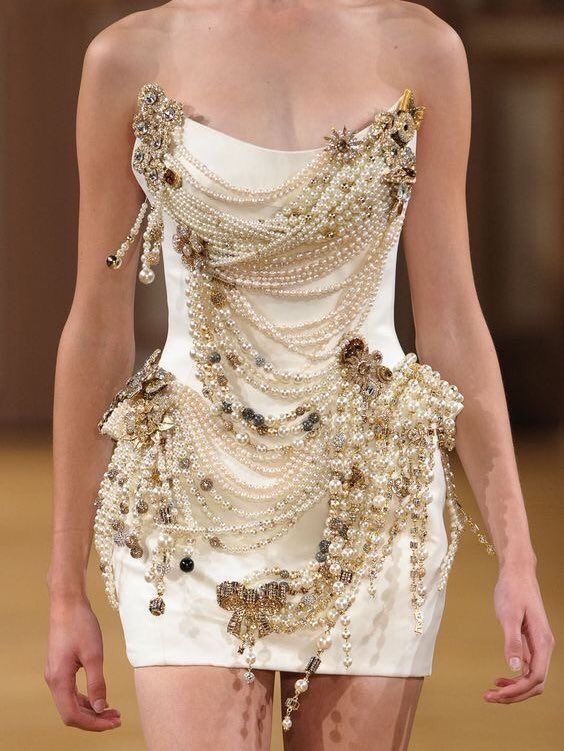 the image shows a model wearing a short, white strapless dress heavily embellished with strands of pearls and other beads. the jewelry is concentrated around the neckline, shoulders, and hip area, creating a cascading effect. the dress appears to be a highfashion or couture piece, suggesting luxury and elegance. the overall impression is ornate and visually striking.