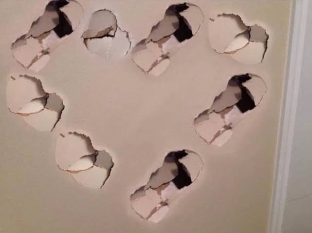 the image shows a wall with multiple holes in it, arranged in the shape of a heart. the damage appears to be recent and significant, exposing the wall's interior. the holes are jagged and rough.
