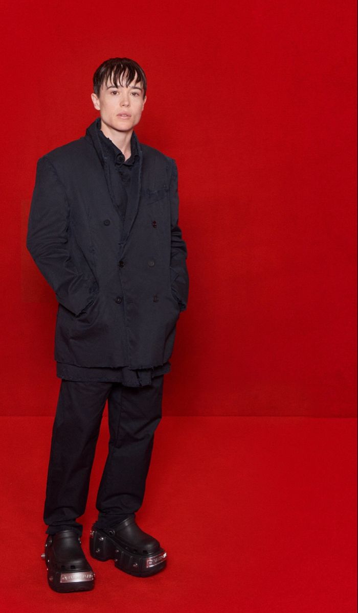a young man stands against a bright red background, wearing a stylish black doublebreasted coat, black pants, and chunky black platform clogs with metallic accents. he is posed in a slightly relaxed stance, his hands in his pockets. his short dark hair is wet, and his expression is serious and somewhat aloof. the overall image is sharp and welllit, emphasizing the fashion elements of the clothing and accessories.